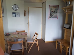 Quarto compacto com uma mesa de jantar de madeira, quatro cadeiras, um relógio de parede, um cartaz e um micro-ondas numa prateleira.