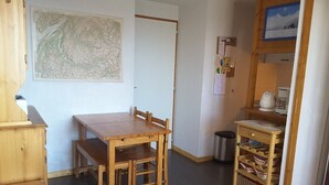 Et lille, hyggeligt værelse med et træbord og stole, et kort på væggen, en lille hylde med service og en skraldespand.