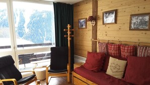Ein gemütliches Zimmer mit Holzboden, einer Couch mit roten Kissen, einem schwarzen Sessel und einem Holztisch.