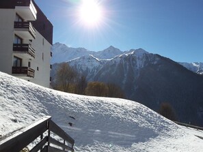 Eine verschneite Berglandschaft mit einem Gebäude, das Balkone und eine hölzerne Bank im Vordergrund zeigt.