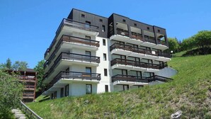 Ein mehrstöckiges Wohngebäude mit Balkonen, eine Treppe führt hinauf, und ein grasbewachsener Hügel ist im Hintergrund zu sehen.