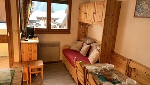 A cozy room with a bed, wooden furniture, a dining table with bowls and cups, and a window with a view of buildings and mountains. - Paradiski ski in ski out vallandry (LANDRY)