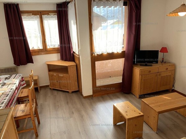 A room with a dining table, chairs, a TV on a wooden cabinet, and a window with curtains.