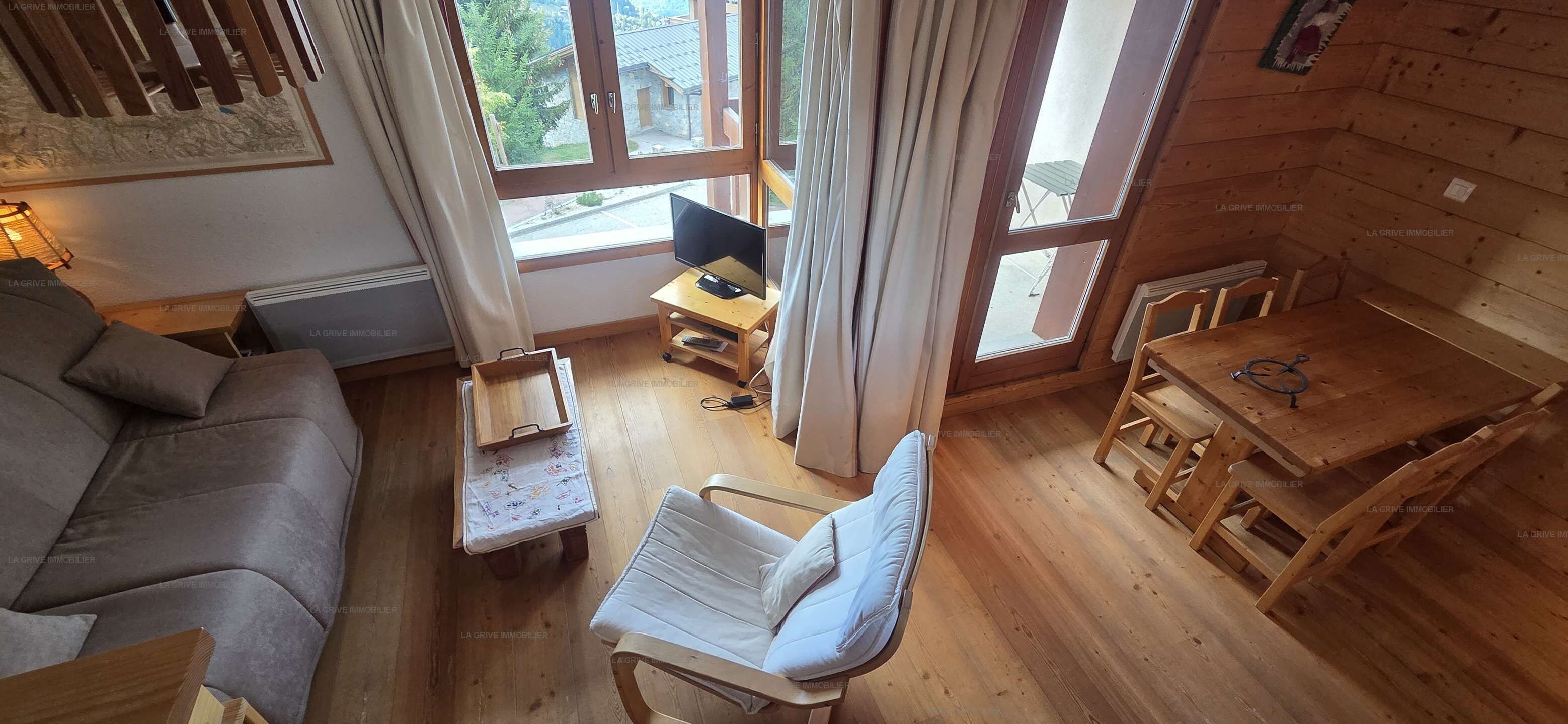 A cozy room with a sofa, a small table with a chair, a TV on a stand, and a window with curtains.