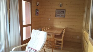 L’intérieur d’un chalet en bois comprend une table à manger, des chaises, un fauteuil blanc rembourré, une fenêtre avec des rideaux et un tableau accroché au mur.