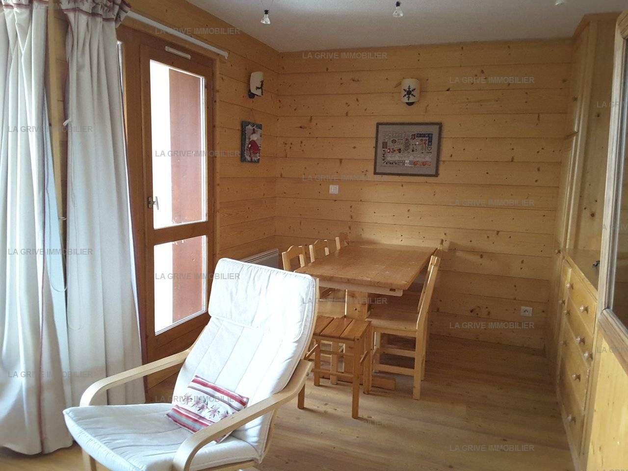 Un intérieur en bois avec une table à manger et des chaises, un fauteuil blanc et une fenêtre avec des rideaux.
