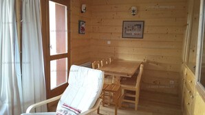 A wooden interior with a dining table and chairs, a white armchair, and a window with curtains.
