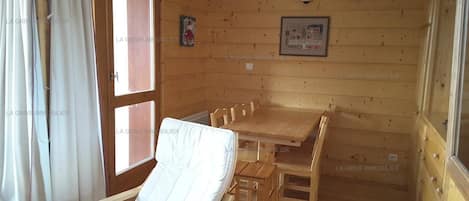 Un intérieur en bois avec une table à manger et des chaises, un fauteuil blanc et une fenêtre avec des rideaux.