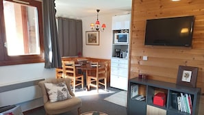 Uma sala de estar acolhedora com um sofá, poltrona e mesa de centro. Uma área de jantar com mesa e cadeiras de madeira é visível ao fundo. Uma TV está fixada na parede e uma estante está encostada na parede.
