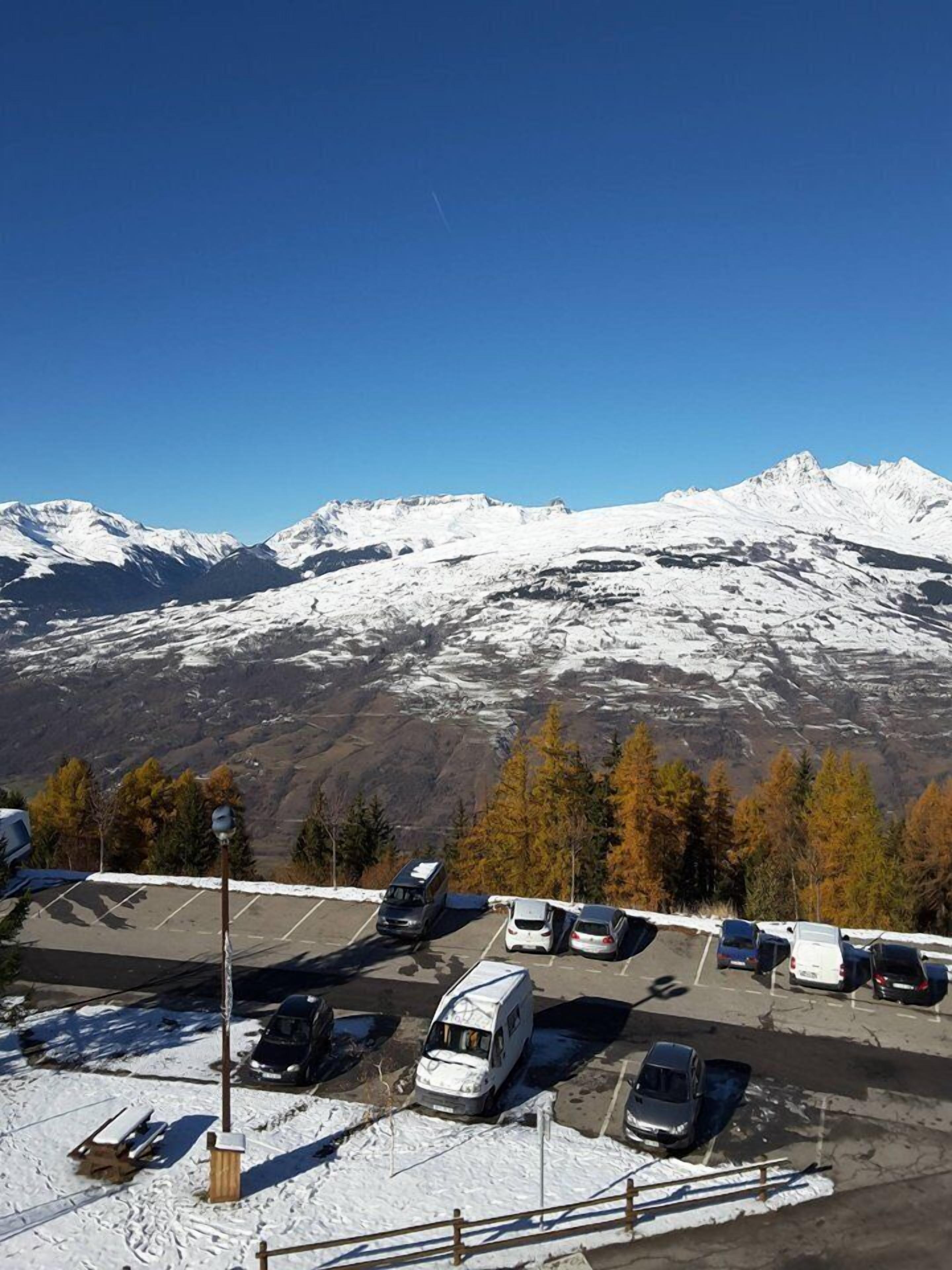 A parking lot with several vehicles, including a white van and a camper van, in front of a mountain range with snow-covered peaks.