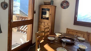 Uma área de refeições com uma mesa de madeira posta para dois, um armário com um relógio e vista para uma paisagem nevada através de uma grande janela.
