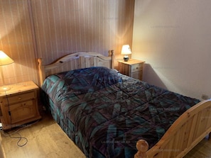 Quarto com cama de madeira, mesa de cabeceira com abajur e mesinha de apoio.