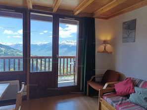 Quarto com teto de madeira, varanda com vista para montanhas, mesa, cadeira e sofá com almofadas.