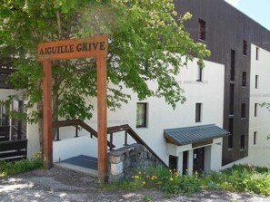 Une enseigne en bois portant l’inscription « AIGUILLE GRIVE » est placée devant un bâtiment à l’architecture moderne, doté d’un escalier et d’un petit espace porche.