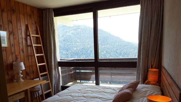 A bedroom with a large bed, a wooden desk, a ladder, and a view of a forested mountain.