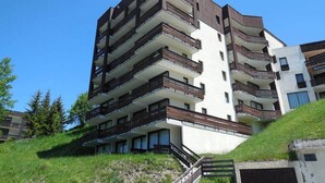 A multi-story residential building with balconies, a staircase, and a grassy hill in the foreground.