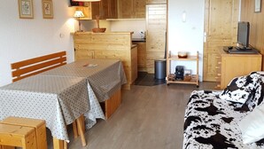 A cozy room with a wooden dining table, a sofa, and a kitchen area with wooden cabinets and a red hanging lamp.