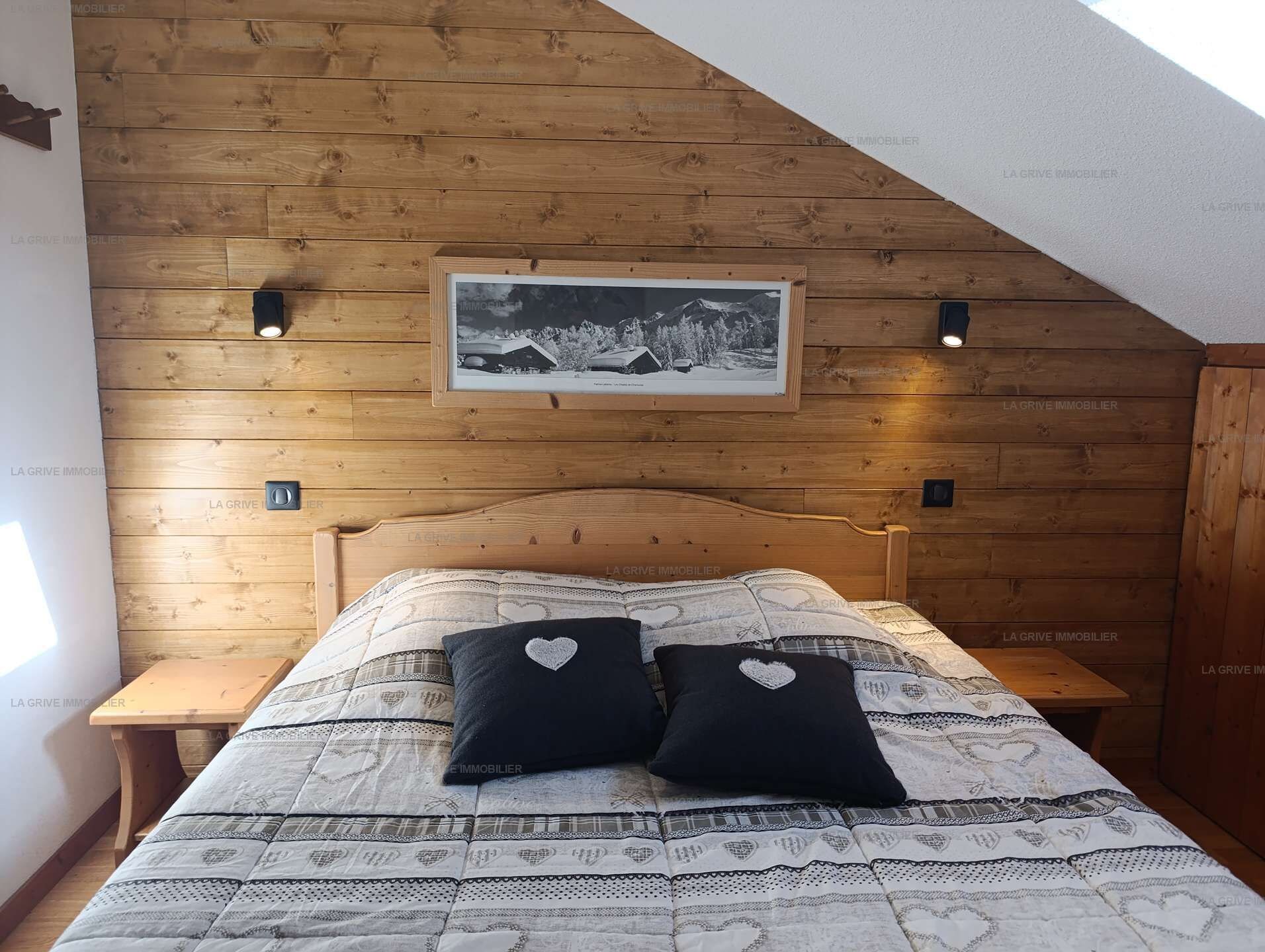 A bedroom with a wooden sloped ceiling, a bed with patterned bedding, two heart-shaped pillows, and a framed picture on the wall.