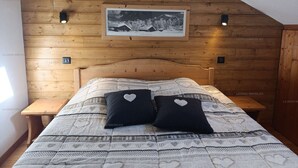 Een slaapkamer met een houten schuine plafond, een bed met beddengoed met een patroon, twee hartvormige kussens en een ingelijst schilderij aan de muur.