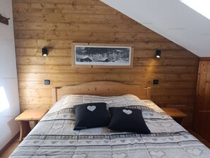 Een slaapkamer met een houten schuine plafond, een bed met beddengoed met een patroon, twee hartvormige kussens en een ingelijst schilderij aan de muur.