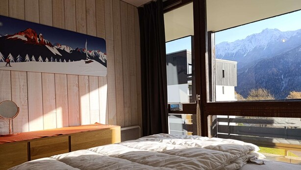 A bedroom with a large bed, a wooden headboard, a bedside table with a mirror, and a mountain view through a large window.