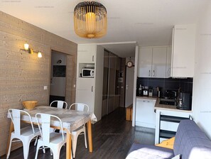 Eine kompakte Küche mit weißen Schränken, einer Mikrowelle und einer Kaffeemaschine. Es gibt einen Essbereich mit Tisch und Stühlen sowie eine Holzwand.