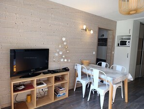 Ein modernes Wohnzimmer mit Flachbildfernseher, Holzregalen, einem Esstisch mit Stühlen und einer Küchenzeile inklusive Mikrowelle.