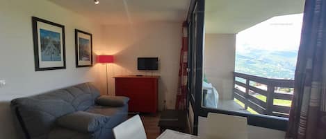 A room with a sofa, a red cabinet, a TV, and a table with papers. There is a window with a view of the outdoors.
