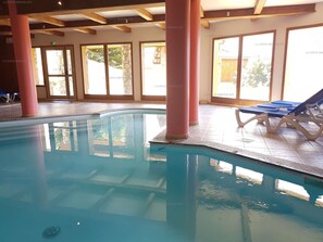 An indoor swimming pool with clear water, surrounded by tiled flooring and large windows.