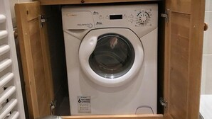 En kompakt, inbyggd tvätt- och torkmaskin med ett träskåp ovanför, en liten papperskorg bredvid och ett klinkergolv.