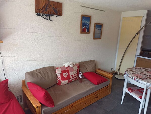 Une pièce chaleureuse avec un canapé en bois, des coussins rouges, une petite table recouverte d’une nappe à carreaux et une lampe fixée au mur.