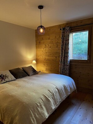 A bedroom with a wooden wall, a bed with white bedding, a hanging light fixture, and a window with curtains.