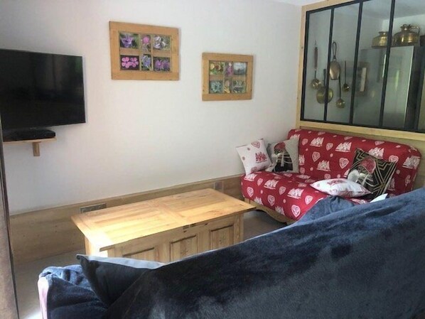 A living room with a wooden table, a red sofa with heart patterns, a TV mounted on the wall, and a cabinet with kitchen utensils.