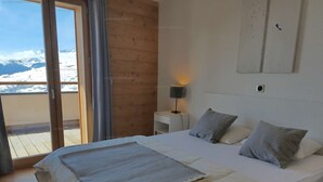 A bedroom with a large bed, a nightstand, a wall-mounted lamp, and a view of snow-capped mountains through a sliding door.