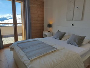A bedroom with a large bed, a nightstand, a wall-mounted lamp, and a view of snow-capped mountains through a sliding door.