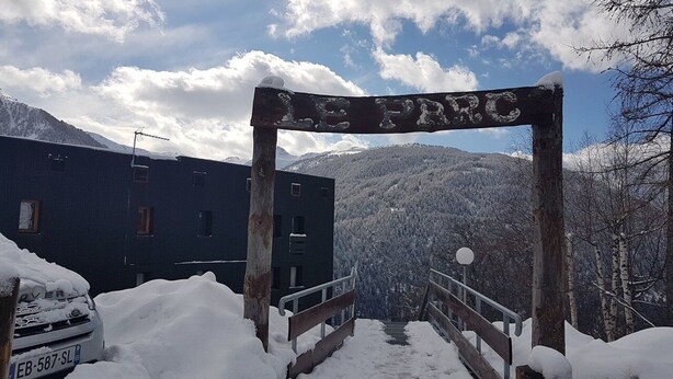 Ein hölzernes Schild mit der Aufschrift 'LE PARC', das zu einem verschneiten Eingang mit Metallgeländer weist.