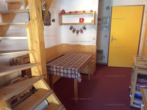 Ein kleines Zimmer mit einem hölzernen Bett, einem Tisch mit kariertem Tuch, einem Regal mit Tassen und einer Tür mit gelbem Griff.