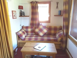 Una habitación acogedora con un sofá de cuadros, una mesita pequeña y una ventana con cortinas.