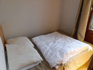 Uma cama de solteiro com um edredom branco e almofadas num quarto com parede lisa e janela com cortinas.