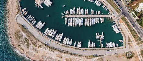 Vista aérea de un puerto deportivo con numerosos barcos amarrados en un arreglo circular, rodeado por una playa de arena y una carretera.