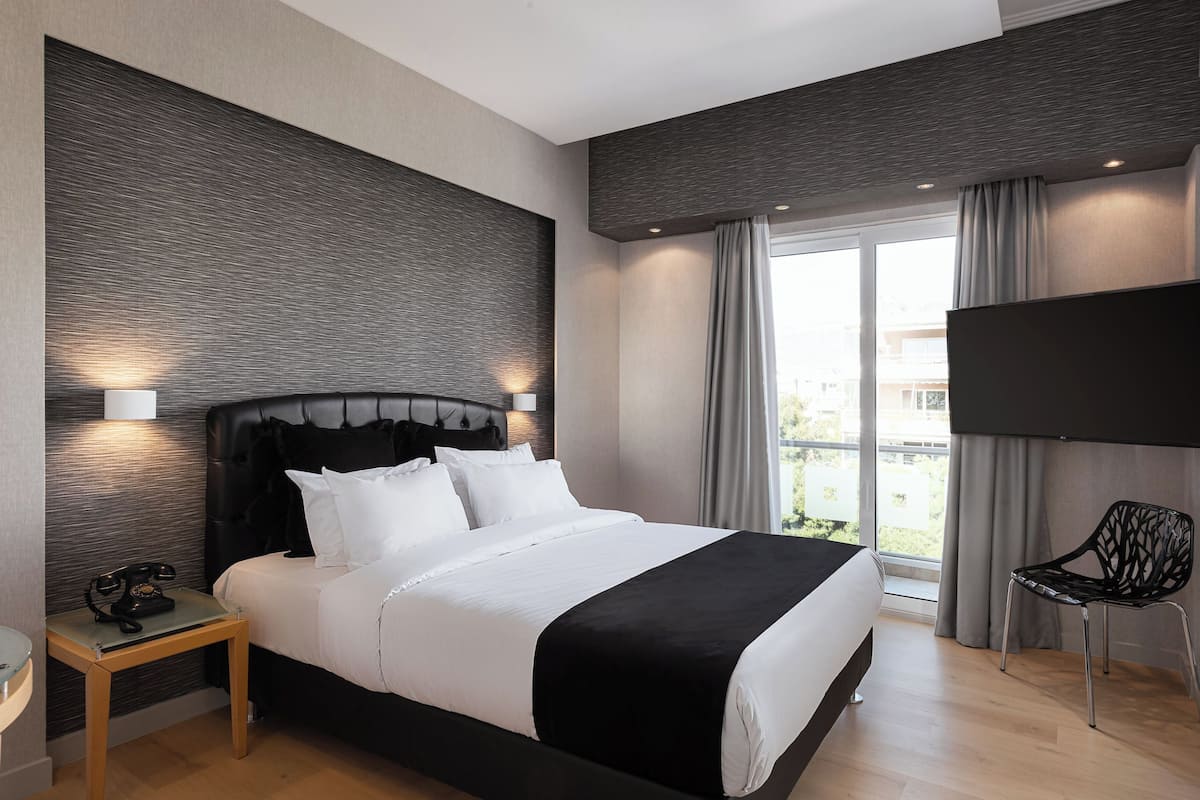 Quarto de hotel moderno com uma cama grande, uma televisão de tela plana, uma mesa de cabeceira de madeira e vista para o exterior por grandes janelas.