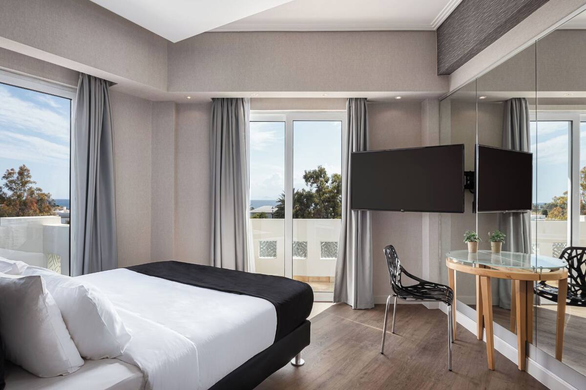 Quarto de hotel moderno com uma cama grande, televisão, mesa de vidro e varanda com vista.