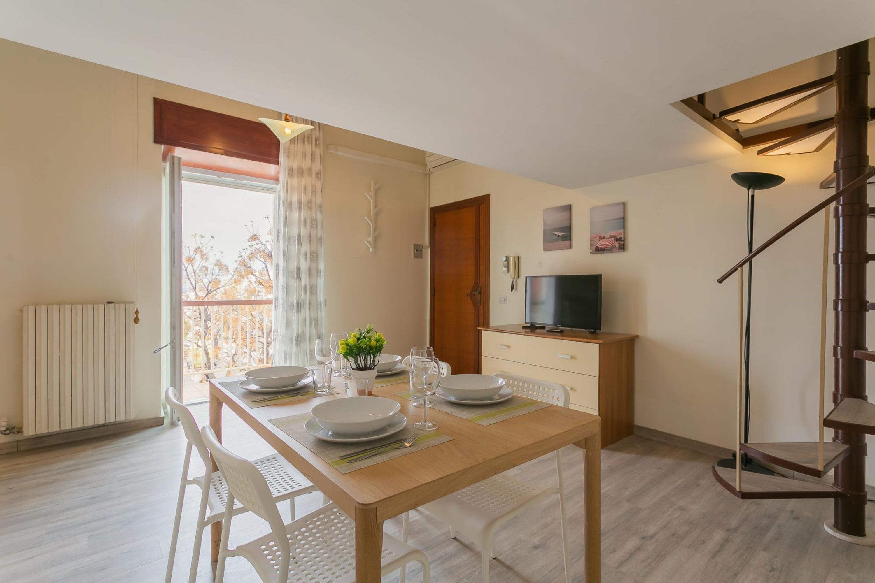 Un comedor con una mesa de madera dispuesta para cuatro personas, sillas blancas, una planta pequeña y un televisor sobre un mueble de madera.