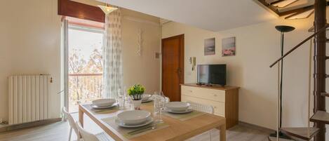 Un comedor con una mesa de madera dispuesta para cuatro personas, sillas blancas, una planta pequeña y un televisor sobre un mueble de madera.
