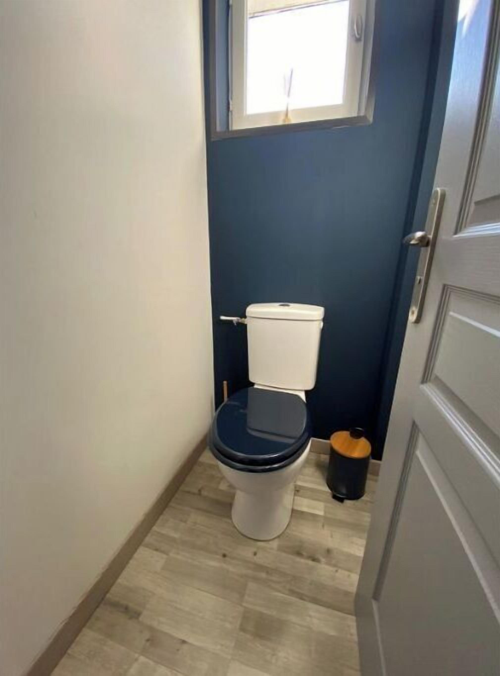 A compact bathroom with a white toilet, a small window above the toilet, and a wooden floor.