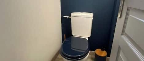 A compact bathroom with a white toilet, a small window above the toilet, and a wooden floor.