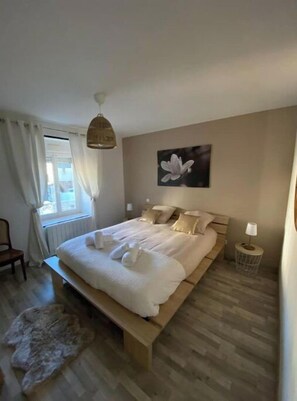 Une chambre à coucher avec un grand lit, un banc, une petite table avec une lampe, un tableau et une fenêtre avec des rideaux.