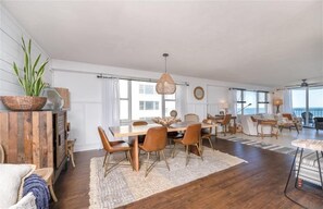 A spacious living area with a wooden dining table, multiple chairs, a large plant, and a ceiling fan.