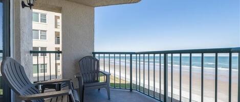 A balcony with two chairs and a table, overlooking a beach with a metal railing.
