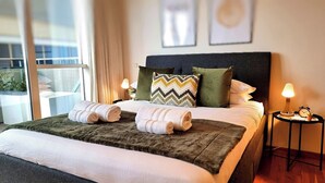 A neatly made bed with two rolled towels, a green throw blanket, and a variety of pillows including a chevron-patterned one. There are two bedside lamps and a small table with a clock and a plant.